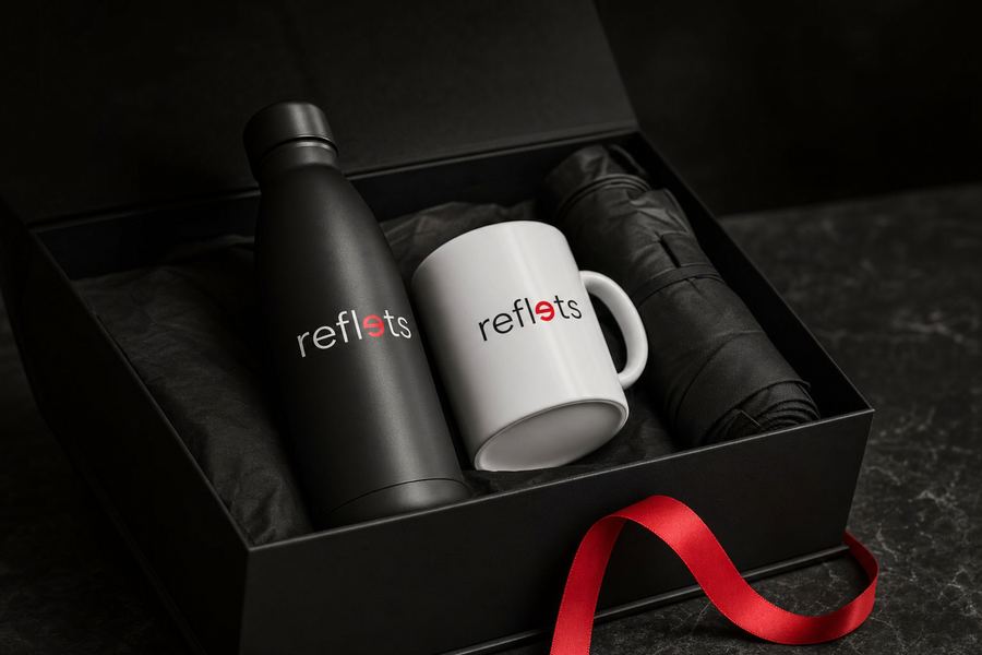Coffret cadeau noir contenant une gourde isotherme noire, un mug blanc et un parapluie pliant marqués du logo Reflets, ruban rouge