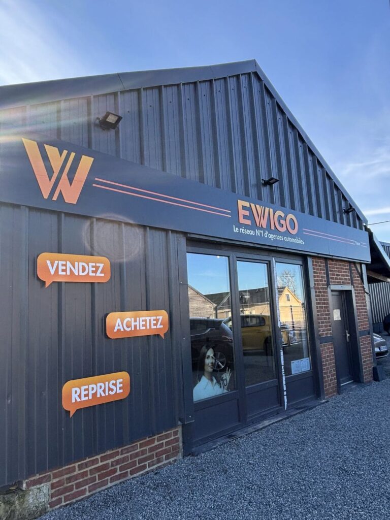 Façade de l'agence automobile Ewigo à Yvetot : bandeau composite gris avec logo et lettrage orange, bulles adhésives Vendez, Achetez, Reprise sur le bardage, enseigne posée par Refletscom