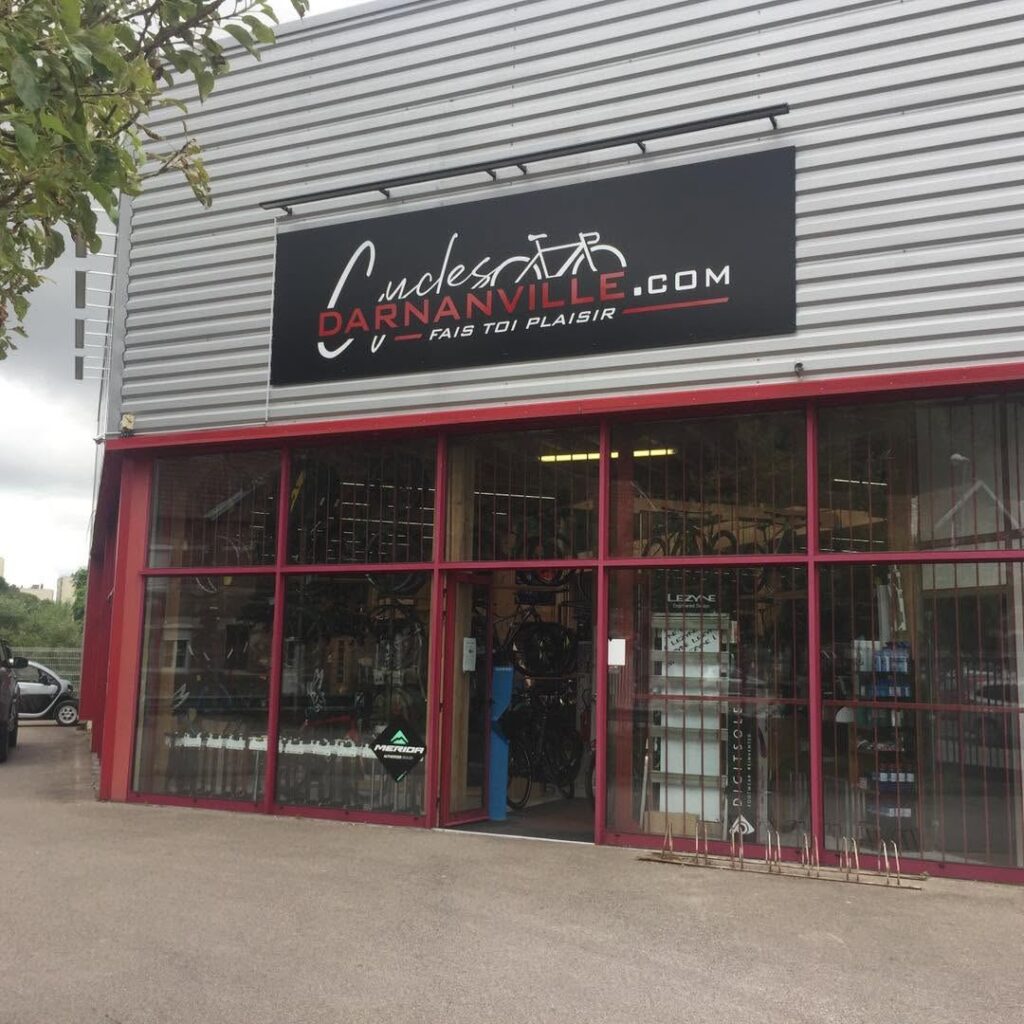 Enseigne bandeau noire avec logo et lettrage adhésifs sur le bardage métallique gris du magasin Cycles Darnanville, posée par Refletscom