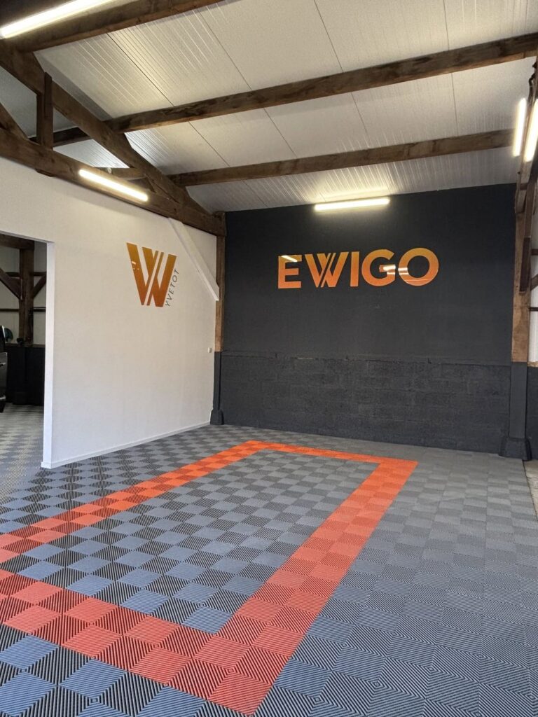 Enseigne intérieure en lettres découpées orange « Ewigo » sur un mur anthracite et logo sur mur blanc dans le showroom de l'agence d'Yvetot, réalisée par Refletscom