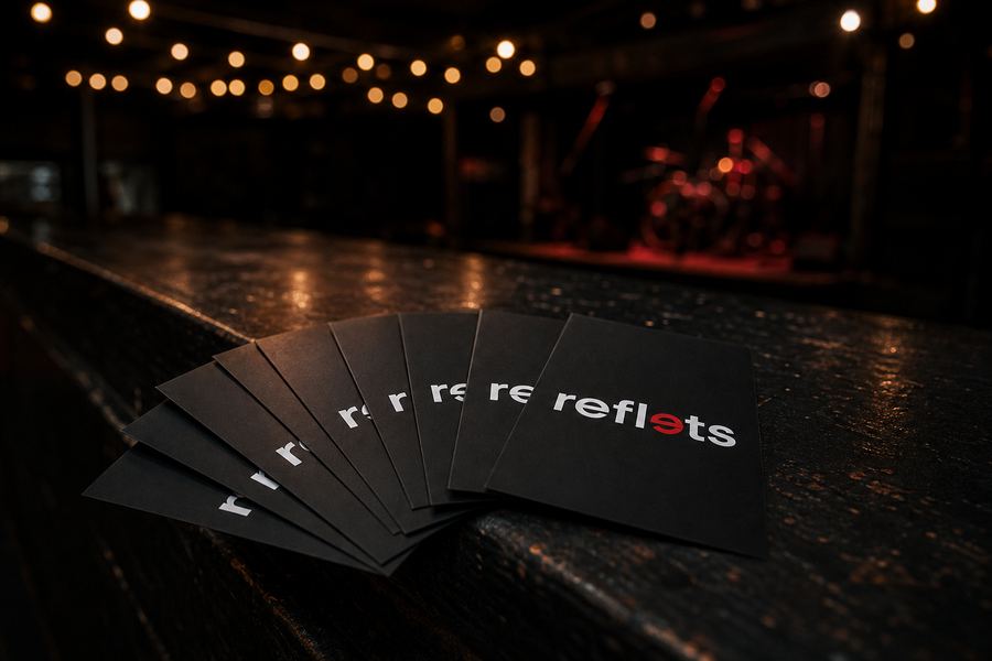 Éventail de flyers A6 noirs au logo Reflets sur le comptoir sombre d'une salle de concert, guirlandes lumineuses et scène floue en arrière-plan