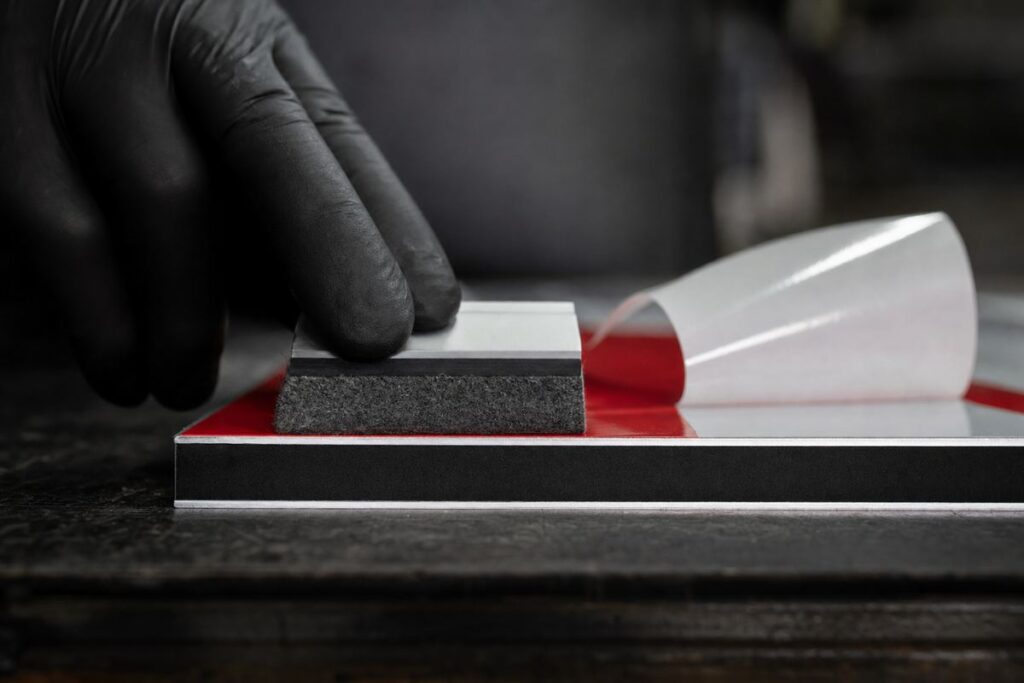 Coupe d'un panneau composite aluminium : deux peaux d'aluminium autour d'une âme noire, avec un film adhésif rouge appliqué à la raclette par une main gantée