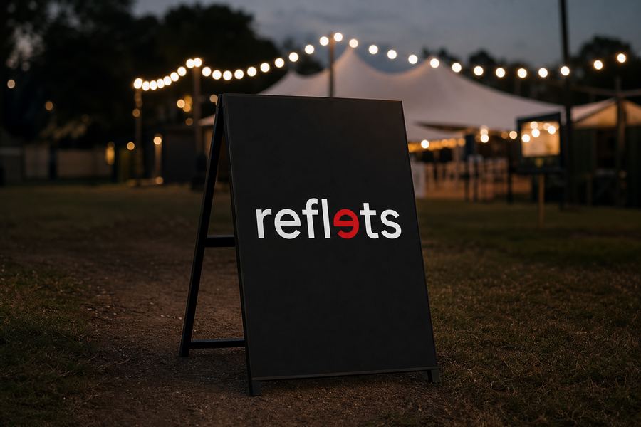 Panneau rigide noir portant le logo Reflets posé sur un chevalet à l'entrée d'un terrain d'événement en plein air, guirlandes lumineuses et chapiteau flous au crépuscule