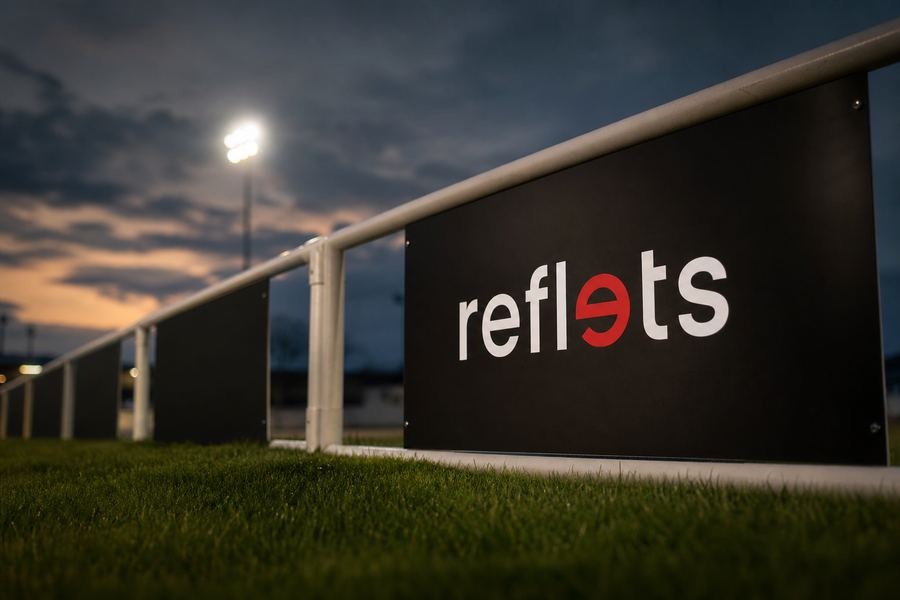 Panneau de sponsor noir portant le logo Reflets fixé sur la main courante d'un terrain de football, pelouse et projecteurs au crépuscule