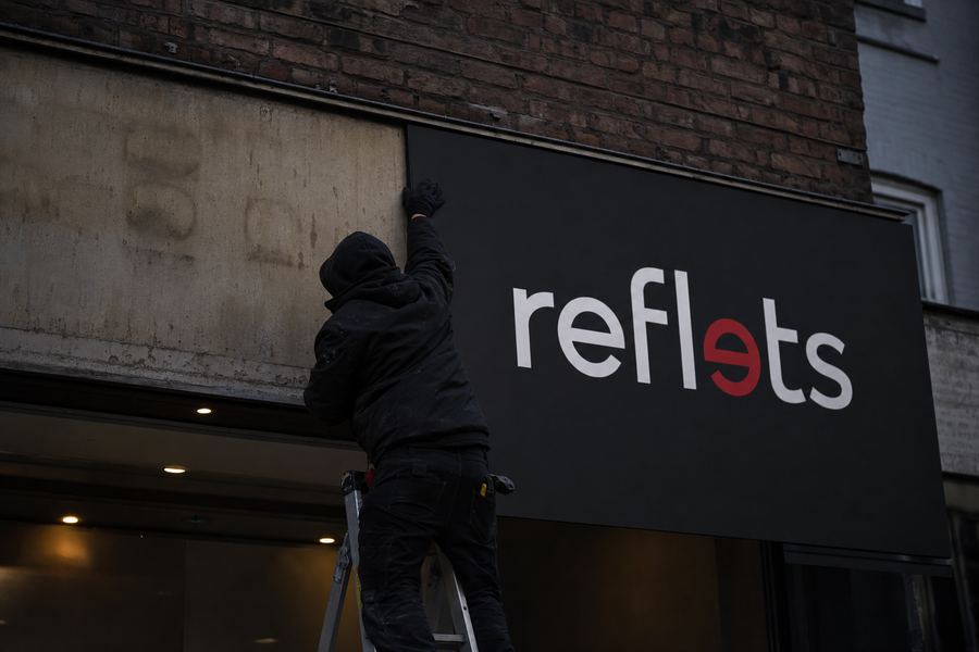 Poseur sur échelle fixant un nouveau panneau composite noir avec le logo Reflets par-dessus une ancienne enseigne de façade décolorée, moitié ancienne moitié neuve, façade en brique