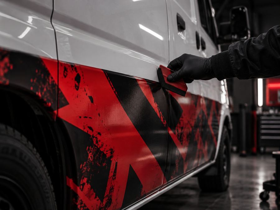 Poseur lissant à la raclette un semi-covering imprimé rouge et noir sur le bas de caisse d’un utilitaire blanc