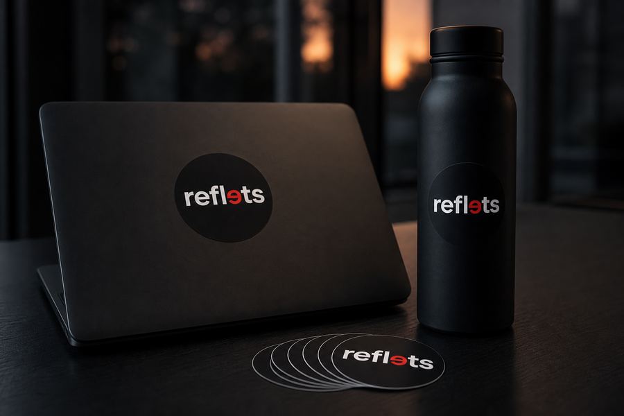Capot d'ordinateur portable noir et gourde noire portant chacun un sticker rond au logo Reflets, pile de stickers identiques sur le bureau sombre