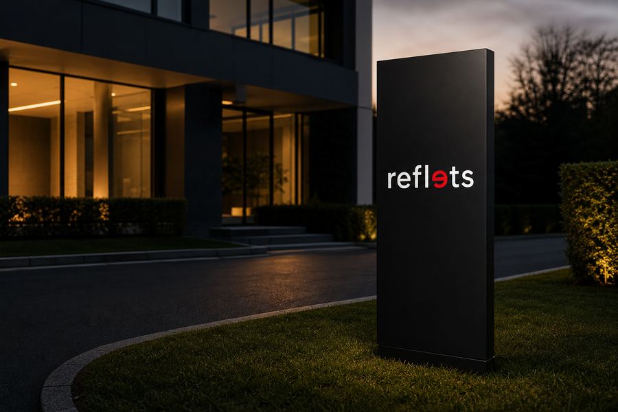 Totem vertical noir portant le logo Reflets planté sur une pelouse à l'entrée d'un bâtiment d'entreprise moderne, au crépuscule